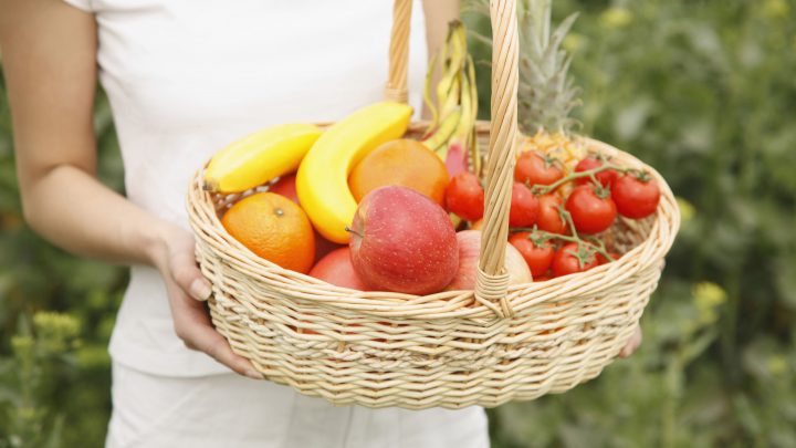 A woman in a white shirt and shorts carries a wicker basket with both hands. In the basket, there is an assortment of produce including oranges, apples, bananas, pineapples, and small tomatoes.