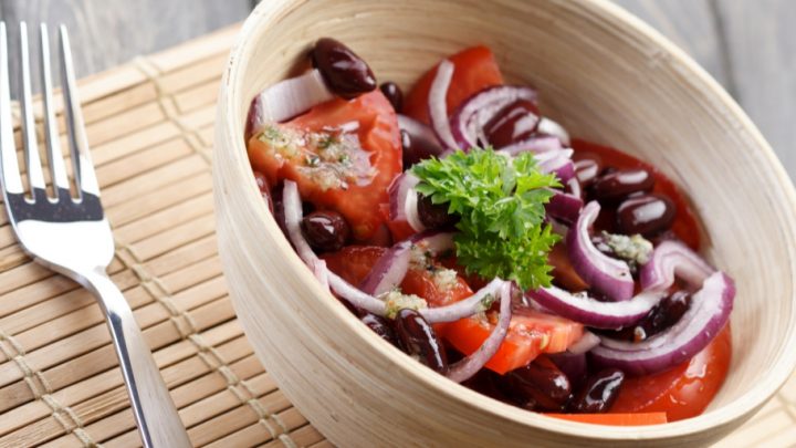 A beige bowl and a stainless steel fork rest on a bamboo mat. In the bowl, there are olives, slices of red onion, and large pieces of chopped tomato.