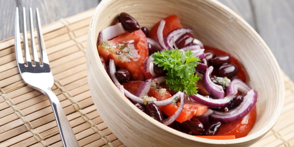 A beige bowl and a stainless steel fork rest on a bamboo mat. In the bowl, there are olives, slices of red onion, and large pieces of chopped tomato.