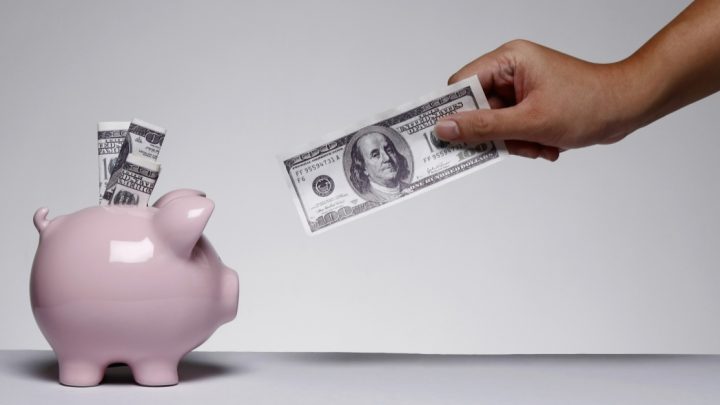 A pink piggy bank has three US 10-dollar bills sticking out of it. A brown hand holding a US 1-dollar bill reaches toward it.