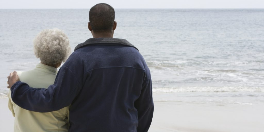 There are two people on the left side of the frame. They are standing with their backs facing the camera and staring out into a large body of water. The man (right) has his left arm around an older woman (left).