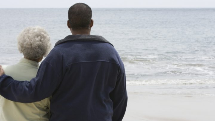 There are two people on the left side of the frame. They are standing with their backs facing the camera and staring out into a large body of water. The man (right) has his left arm around an older woman (left).