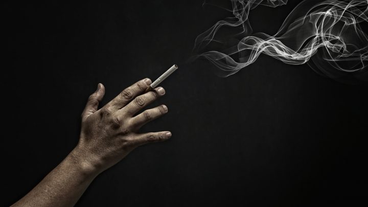 On a black background, a cigarette is being held between the first and middle fingers. A puff of smoke is visible in the top right corner.
