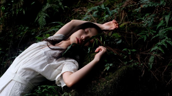 Person in white dress lying on a mossy rock with lush green trees all around them.