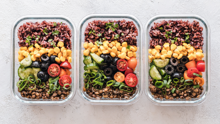 Three glass lock containers containing the same meal of brown rice, corn, cucumber slices, black olives, cherry tomatoes, green onion, and lentils.