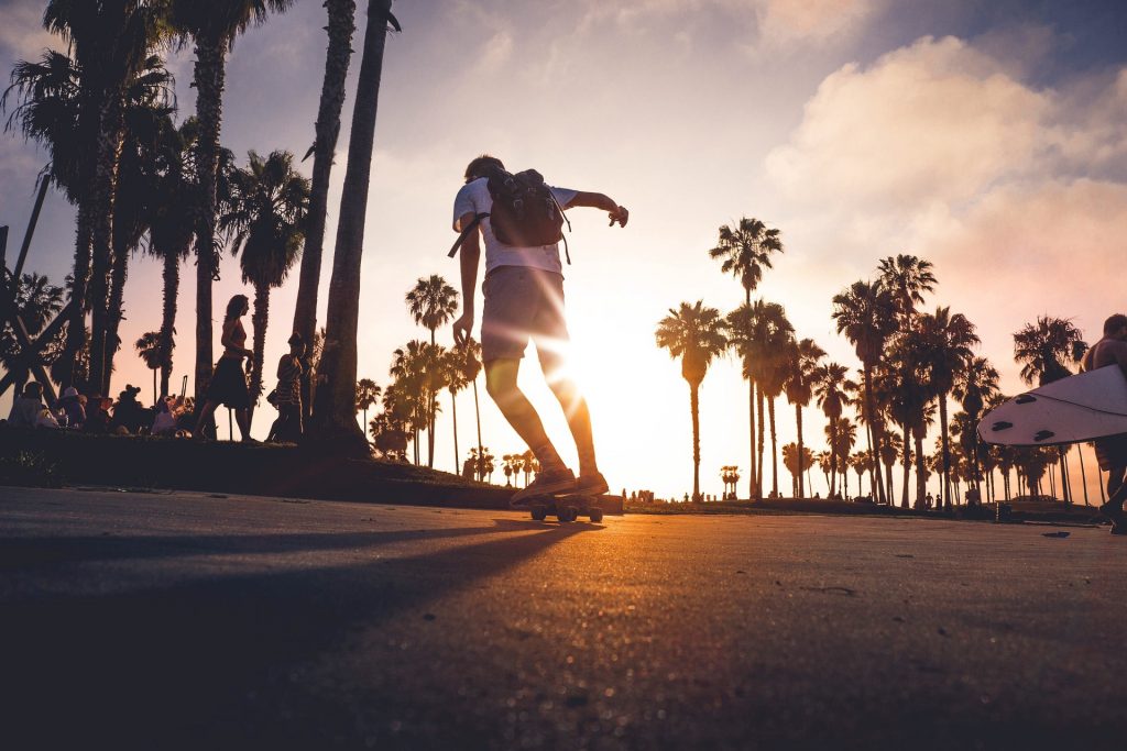 Skateboarder with a backpack on a beachside road with palm trees in the background. Several other people in the background sitting or walking by.