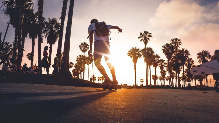 Skateboarder with a backpack on a beachside road with palm trees in the background. Several other people in the background sitting or walking by.